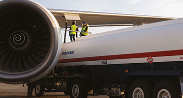 Two technicians in safety vests working on a Boeing aircraft engine, with a fuel truck parked beside it on the tarmac.