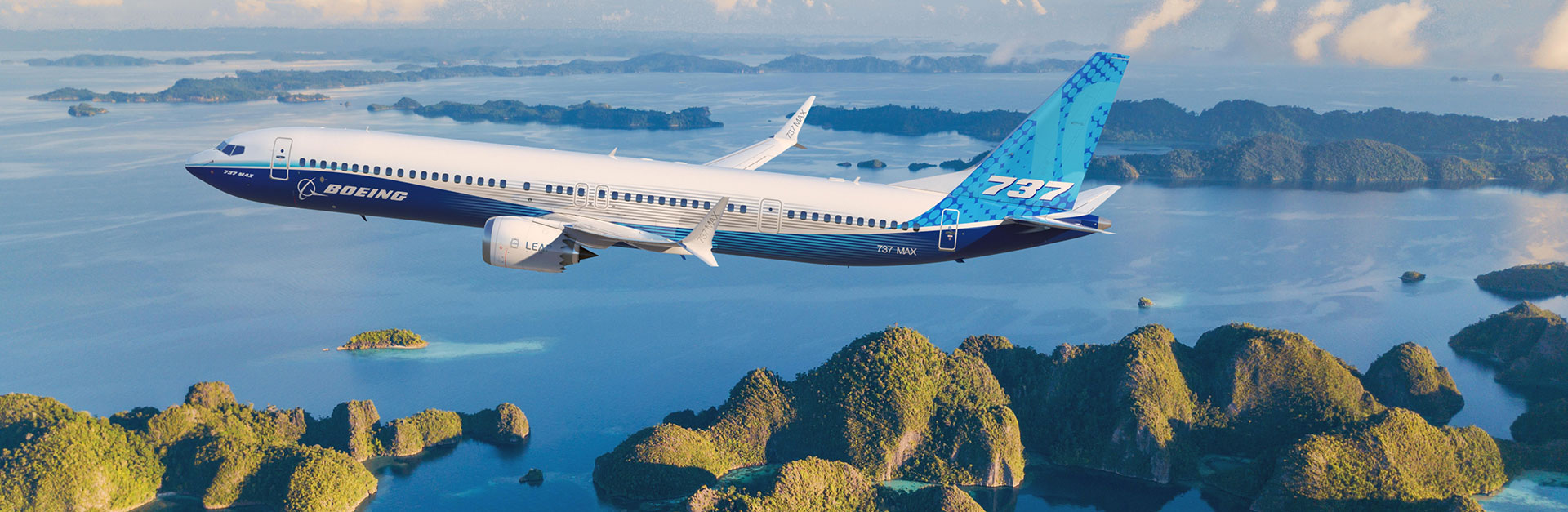 A Boeing 737 MAX 10 flying over a scenic landscape with green forested islands and blue sea below.