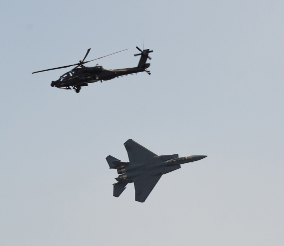 An Apache helicopter flying above an F-15 fighter jet in a gray sky.